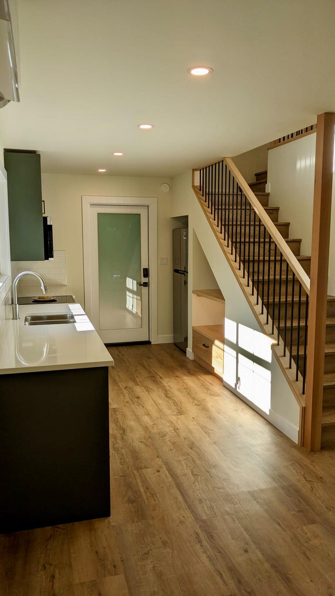 Open-Concept Living Area & Built-In Storage Open-concept interior showing kitchen counter, wood stairwell with built-in under-stair storage nook, and modern front door.