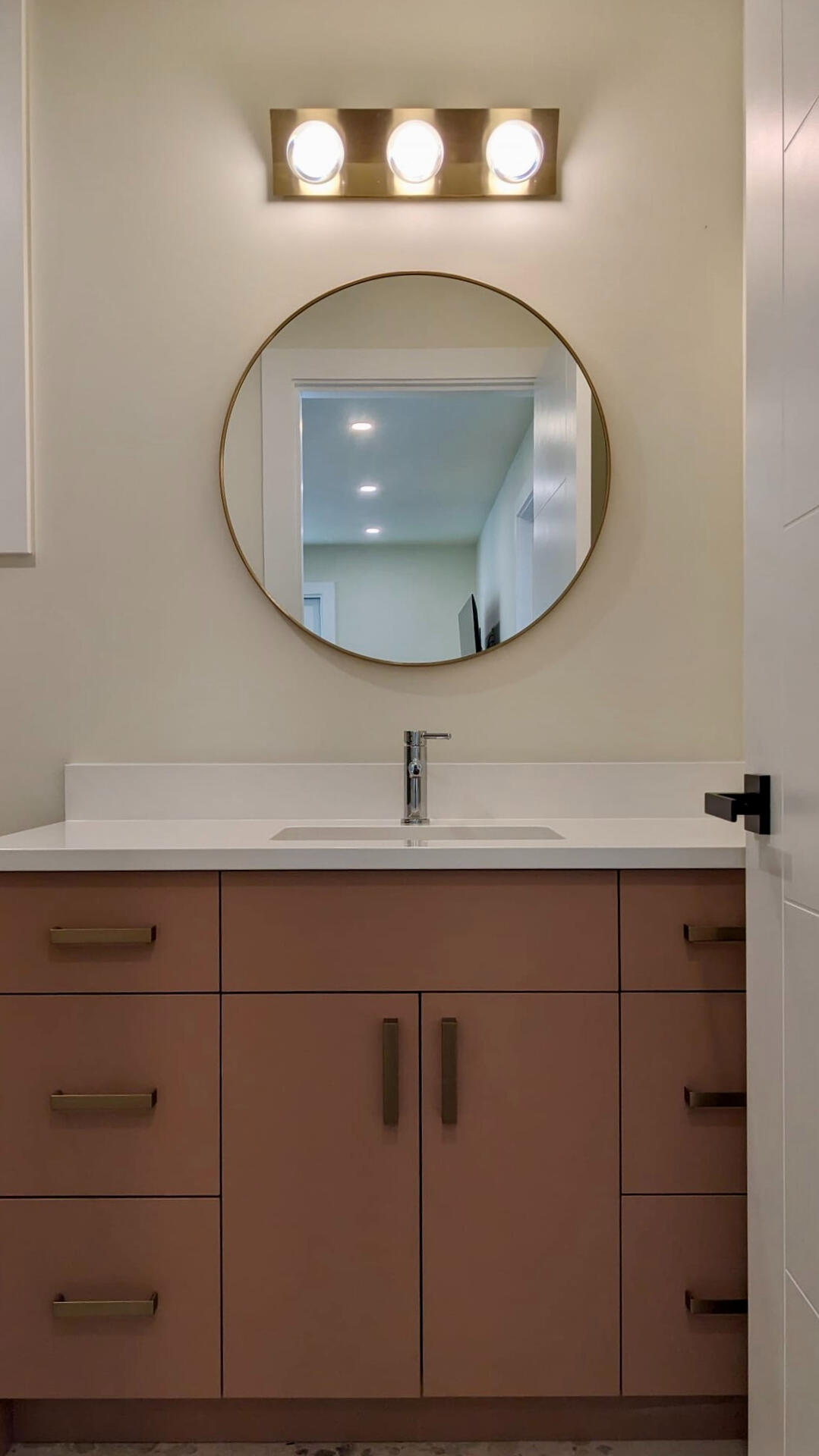 Modern Bathroom Vanity with Brass Accents Terracotta bathroom vanity featuring a white quartz countertop, circular gold frame mirror, and modern three-light vanity bar.