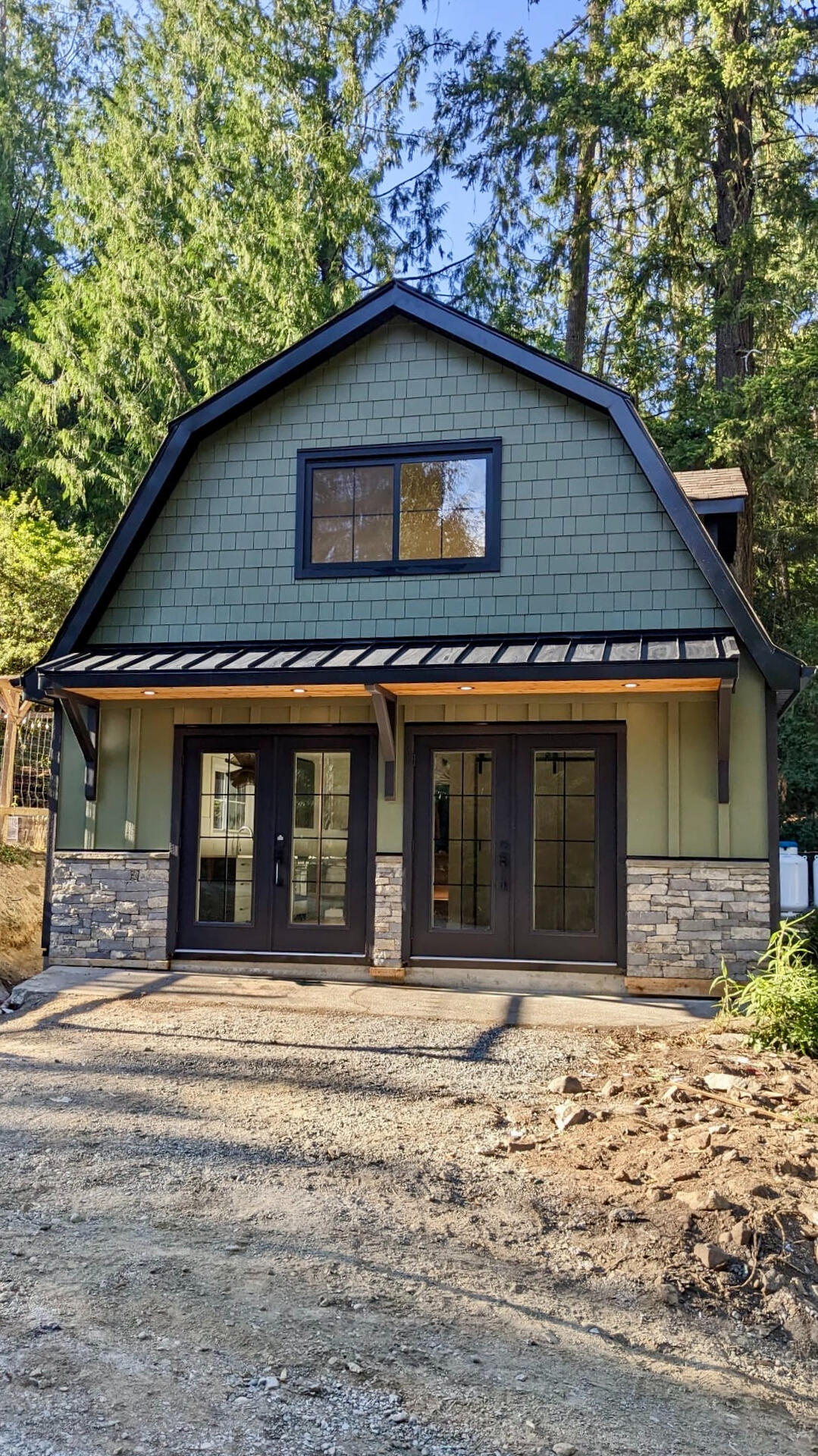 Custom Sage Green Carriage House Exterior Exterior view of a two-story modern carriage home with green vertical siding, stone veneer accents, and glass double doors surrounded by trees.